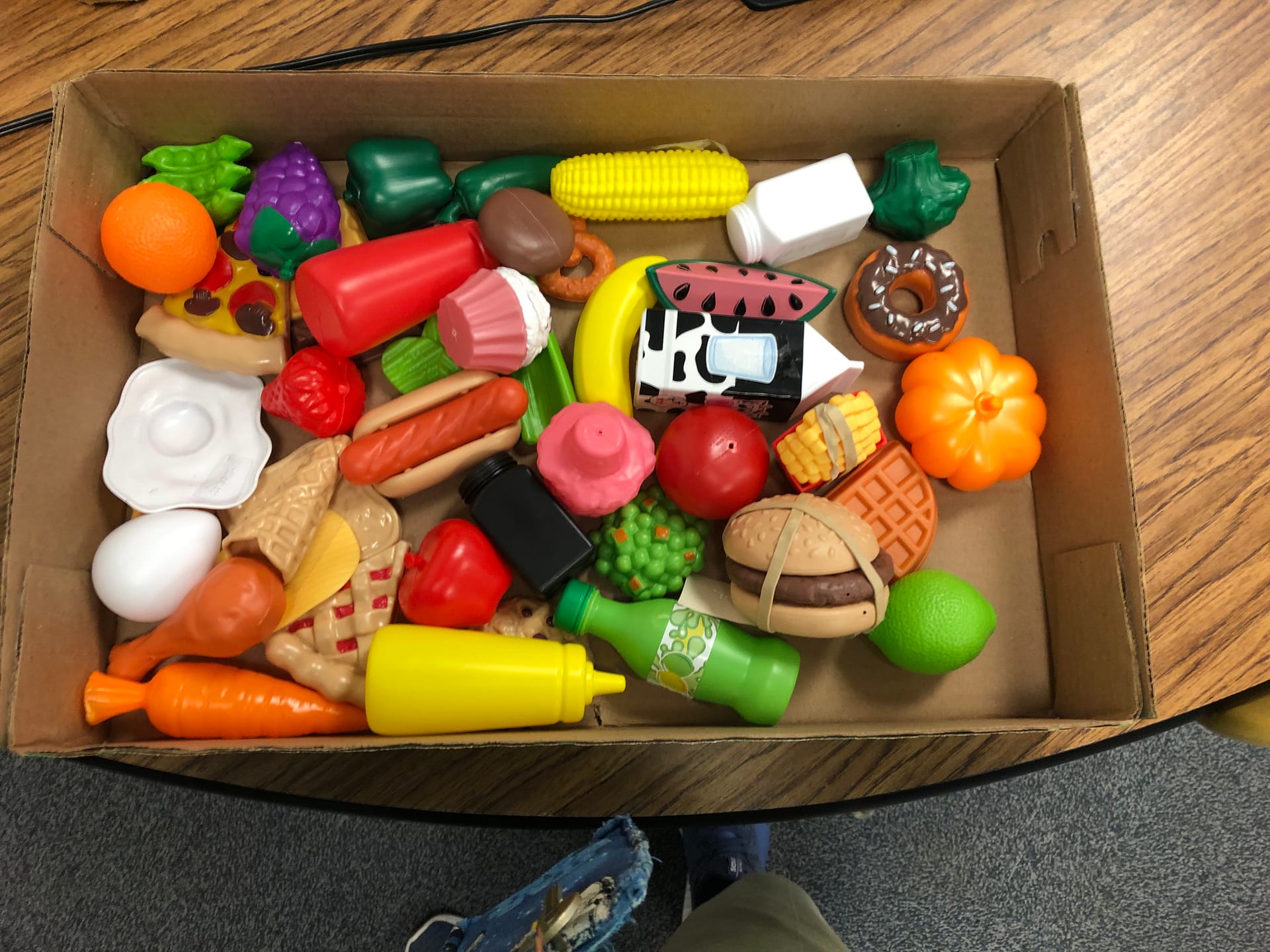 Numerous play food items of different colors in a cardboard box sitting on a table